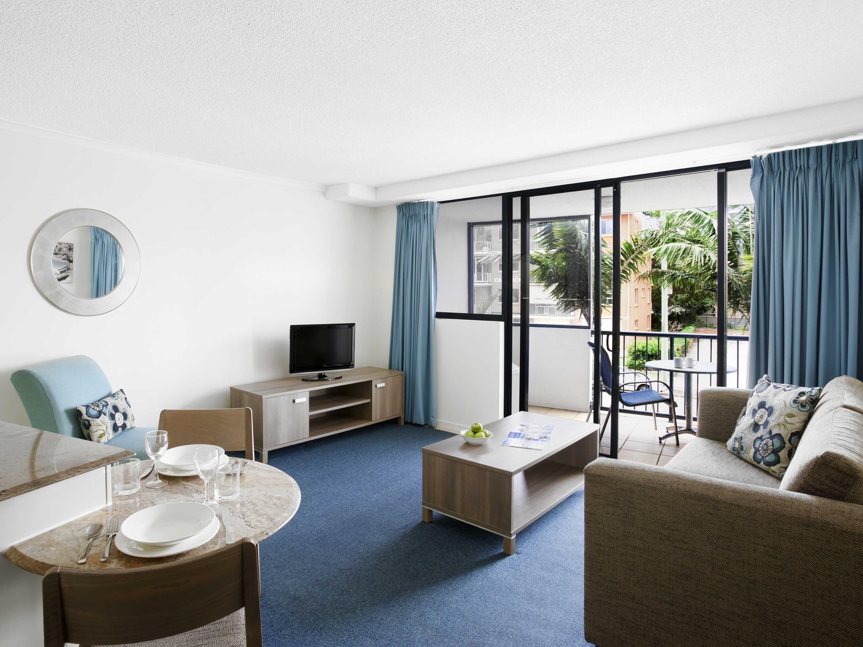 A hotel room with a dining table, a sofa, a TV, and a balcony with a view of greenery.