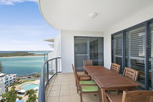 A balcony with a wooden table and chairs, overlooking a pool and beach.