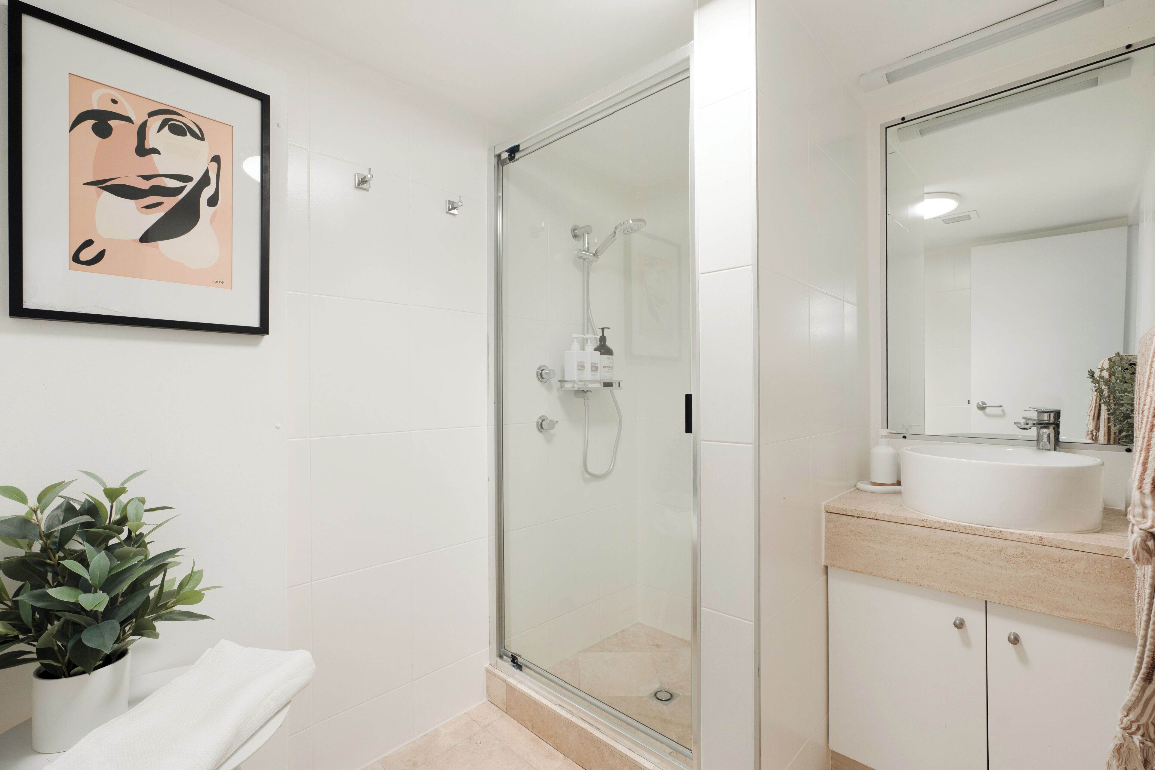A bathroom with a glass-enclosed shower, a sink with a mirror, and a framed artwork on the wall.
