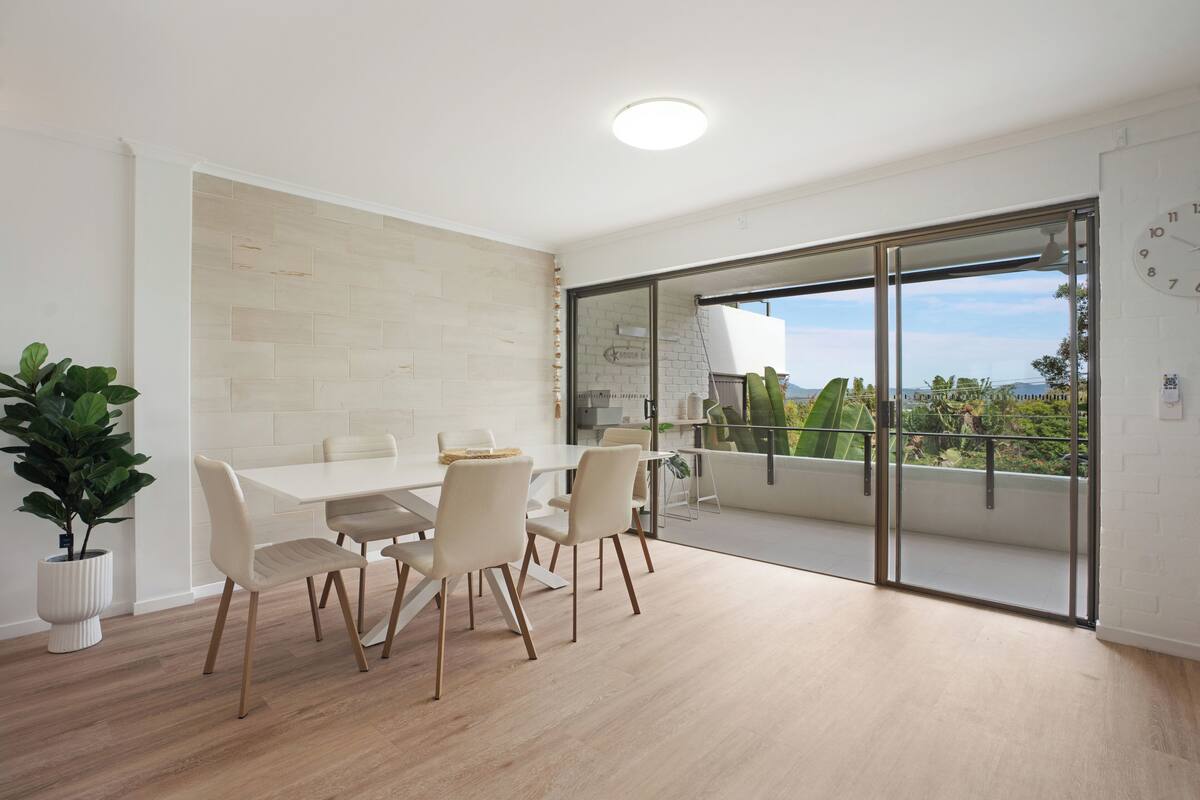 A modern dining area with a white table, four chairs, a potted plant, and a view through a glass door.