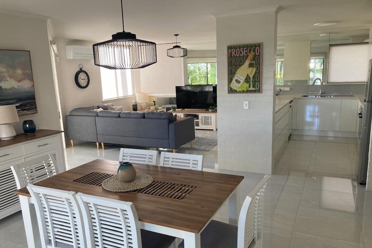 A modern kitchen with white cabinets, a dining area with a wooden table and white chairs, a gray sofa, and a large window.