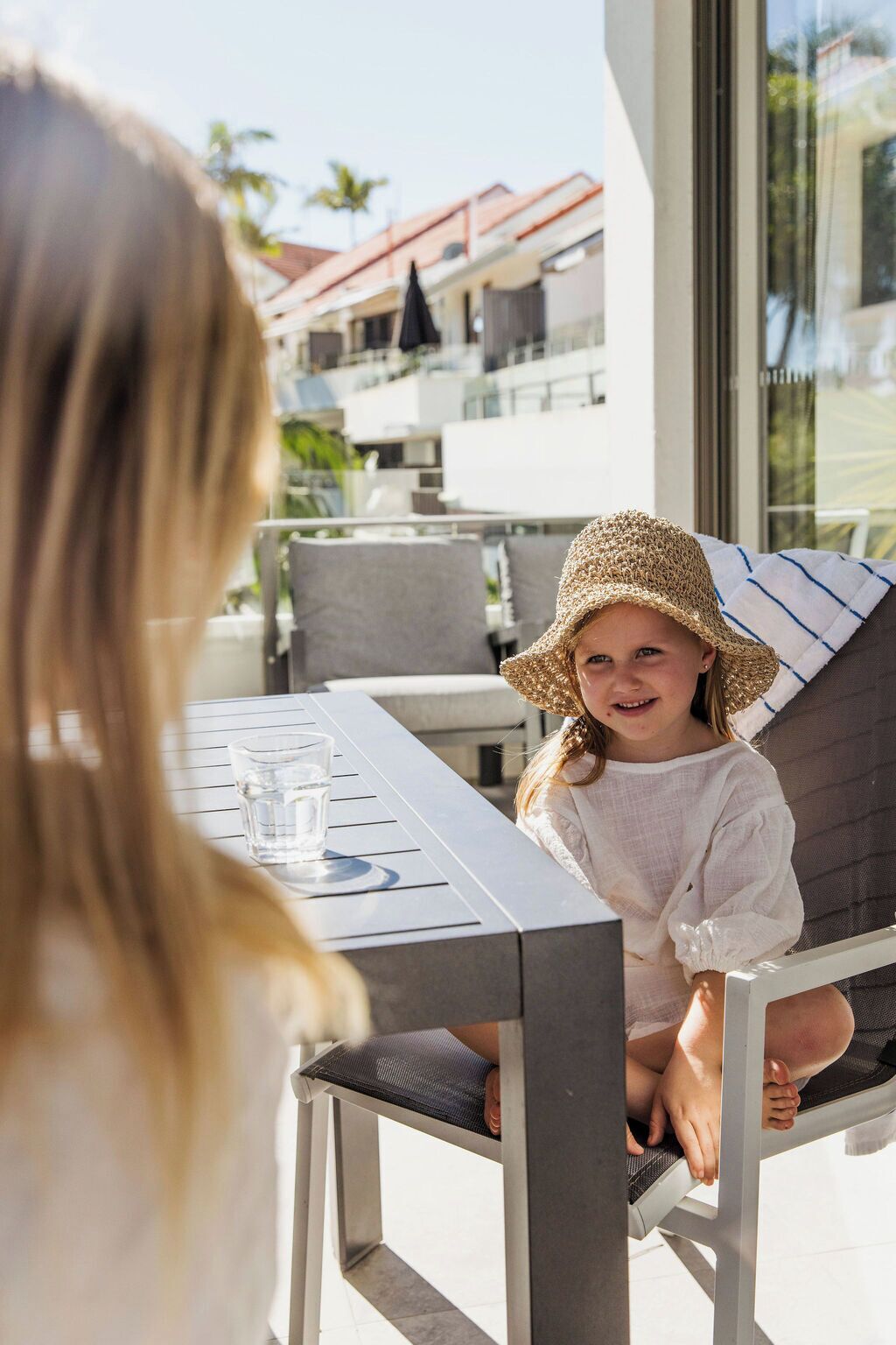 Une personne est assise à une table en plein air, un verre d’eau à portée de main, et elle a devant elle une vue sur un quartier résidentiel avec des bâtiments modernes et un ciel dégagé.