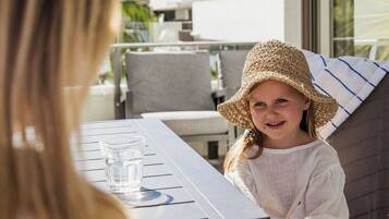 Une personne est assise à une table en plein air, un verre d’eau à portée de main, et elle a devant elle une vue sur un quartier résidentiel avec des bâtiments modernes et un ciel dégagé.