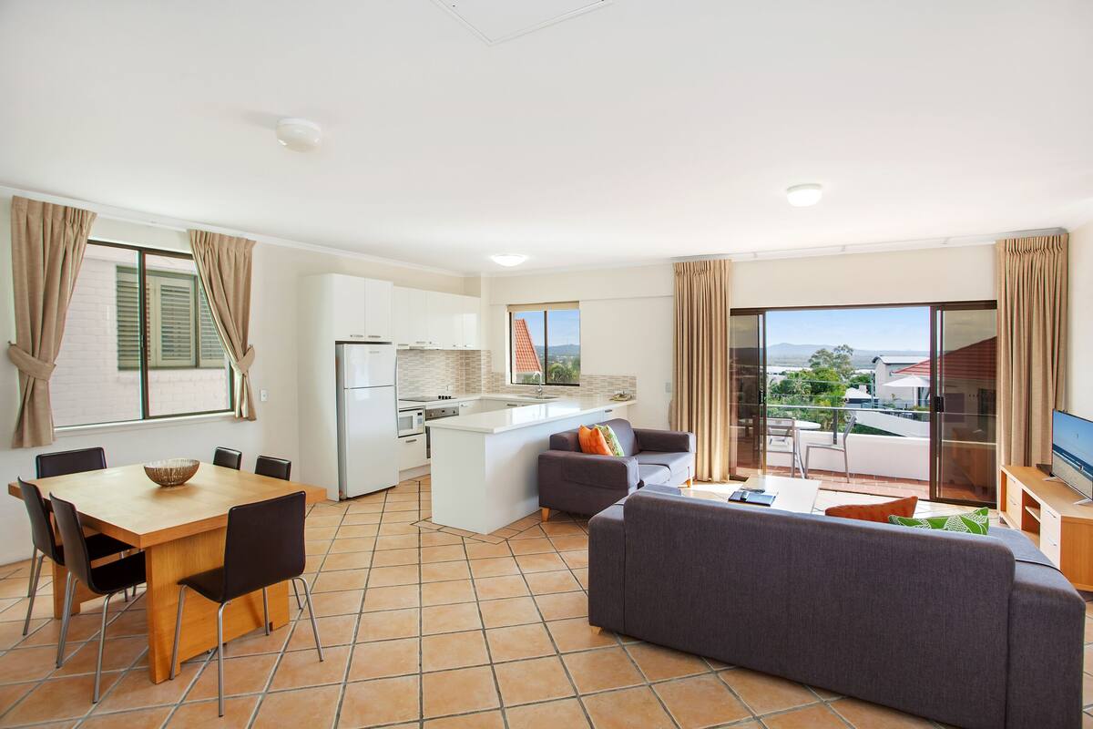 A modern apartment with a kitchen, dining area, and living room. It features a large window with a view, a balcony, and a flat-screen TV.