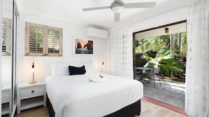 Quarto com cama, mesas de cabeceira, ventilador de teto, janela com vista para o verde e porta que dá para área externa.