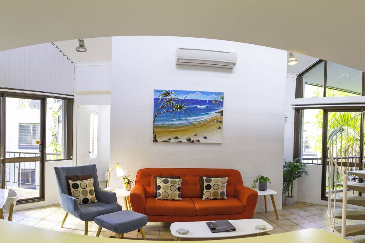 A modern living room with a sofa, armchair, and coffee table, a painting of a beach scene, and a spiral staircase.
