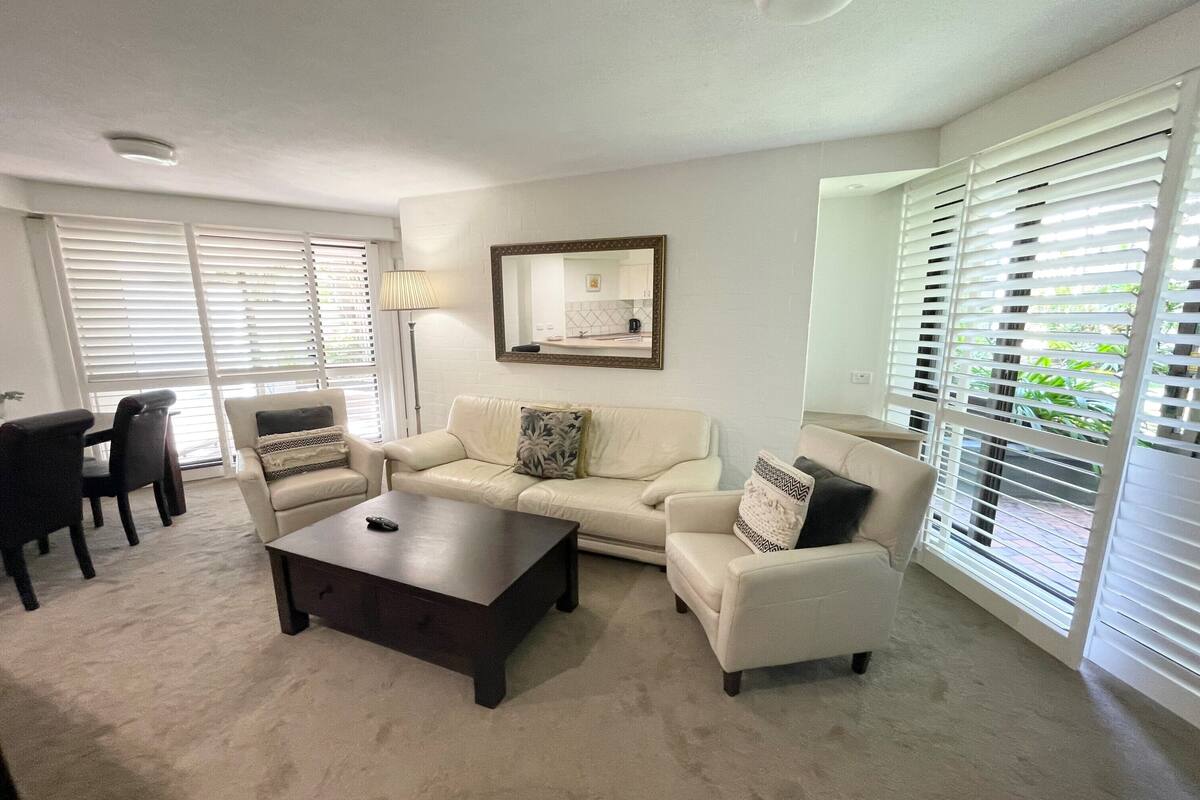 A living room with a beige carpet, a dark wooden coffee table, and a beige sofa with patterned cushions. There is a dining area with a dark wooden table and chairs, and a large window with blinds.