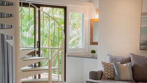 Un interior moderno con una escalera que lleva a un balcón, un sofá con cojines estampados y vistas de abundante vegetación.