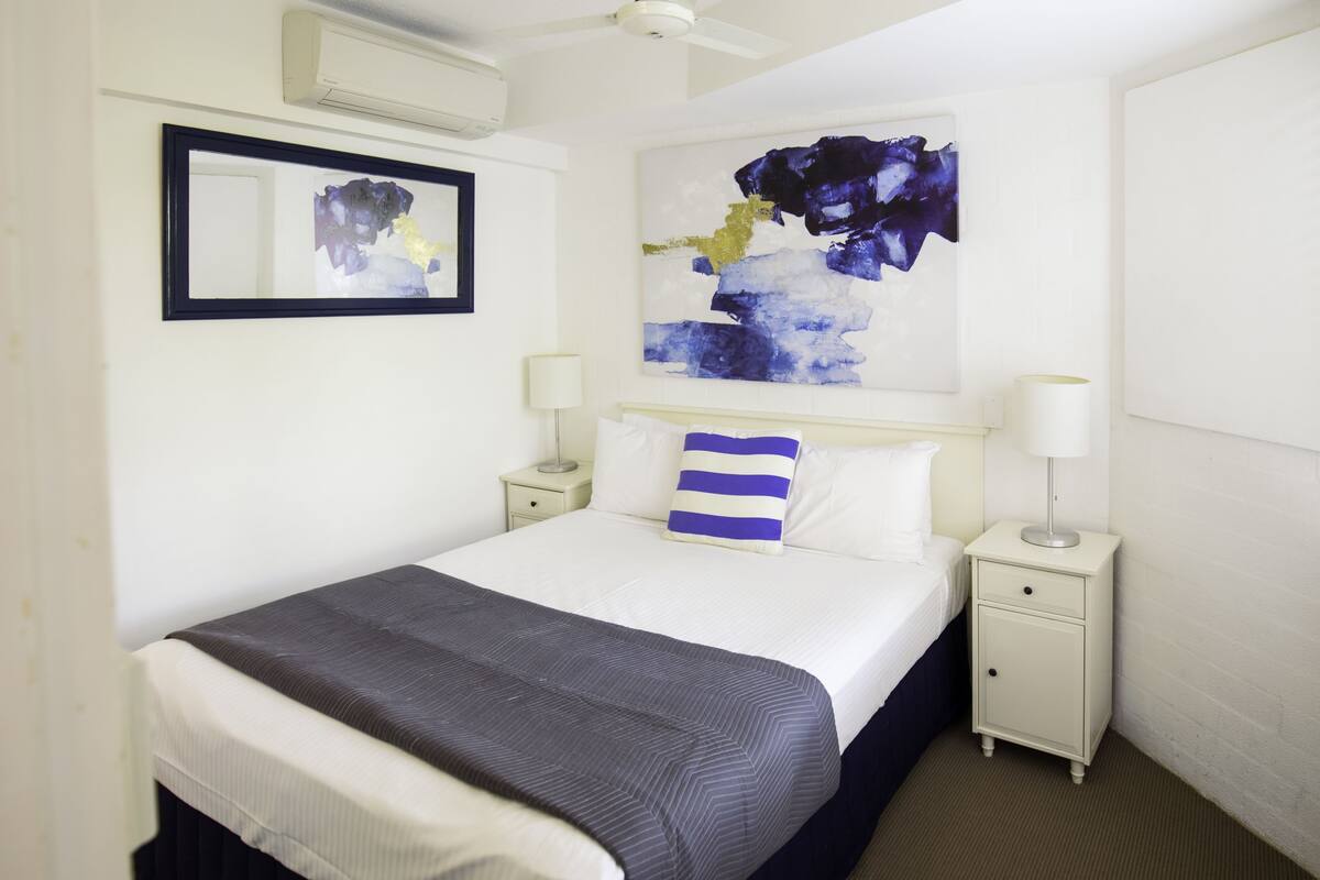 A hotel room with a bed, two bedside tables, a nightstand, a ceiling fan, a window with blinds, and a painting on the wall.