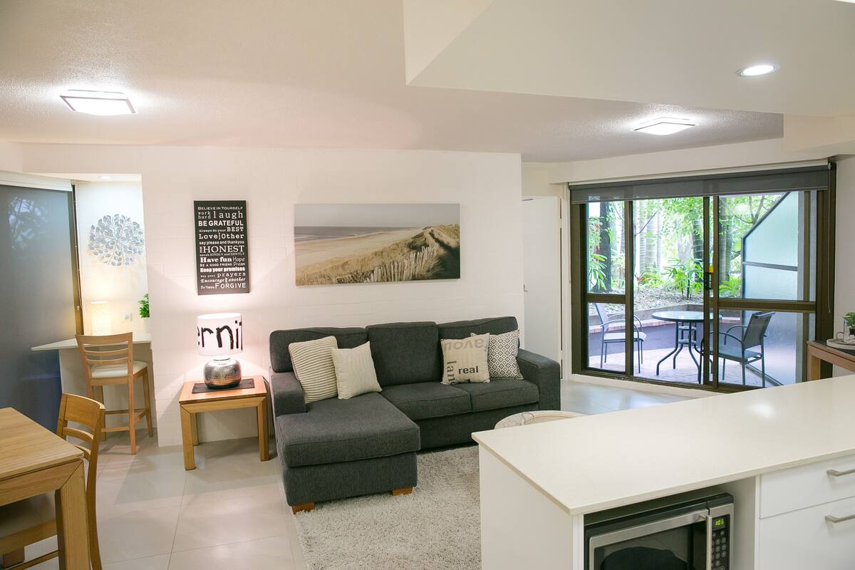 A modern living room with a grey sectional sofa, a wooden dining table, and a kitchen area with a microwave.