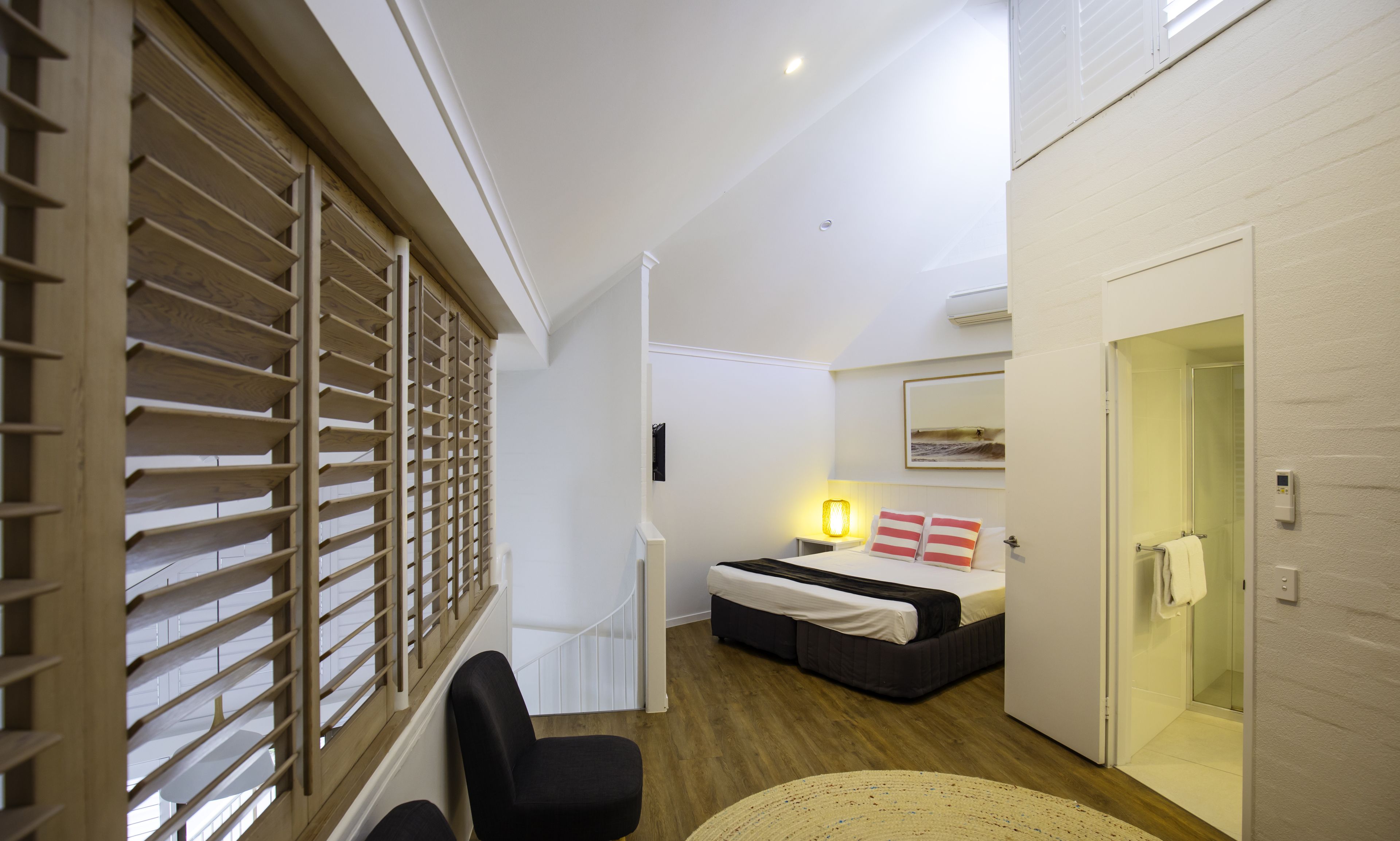 Une chambre d’hôtel comprenant un lit, une table de chevet avec une lampe, une fenêtre avec des stores, une porte et une salle de bain avec une douche.