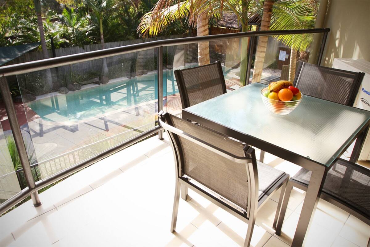 A balcony with a glass table and chairs, overlooking a swimming pool and lush greenery.