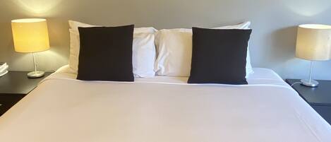 A hotel room with a bed, two black pillows, a white bedspread, and a painting of a wave on the wall.