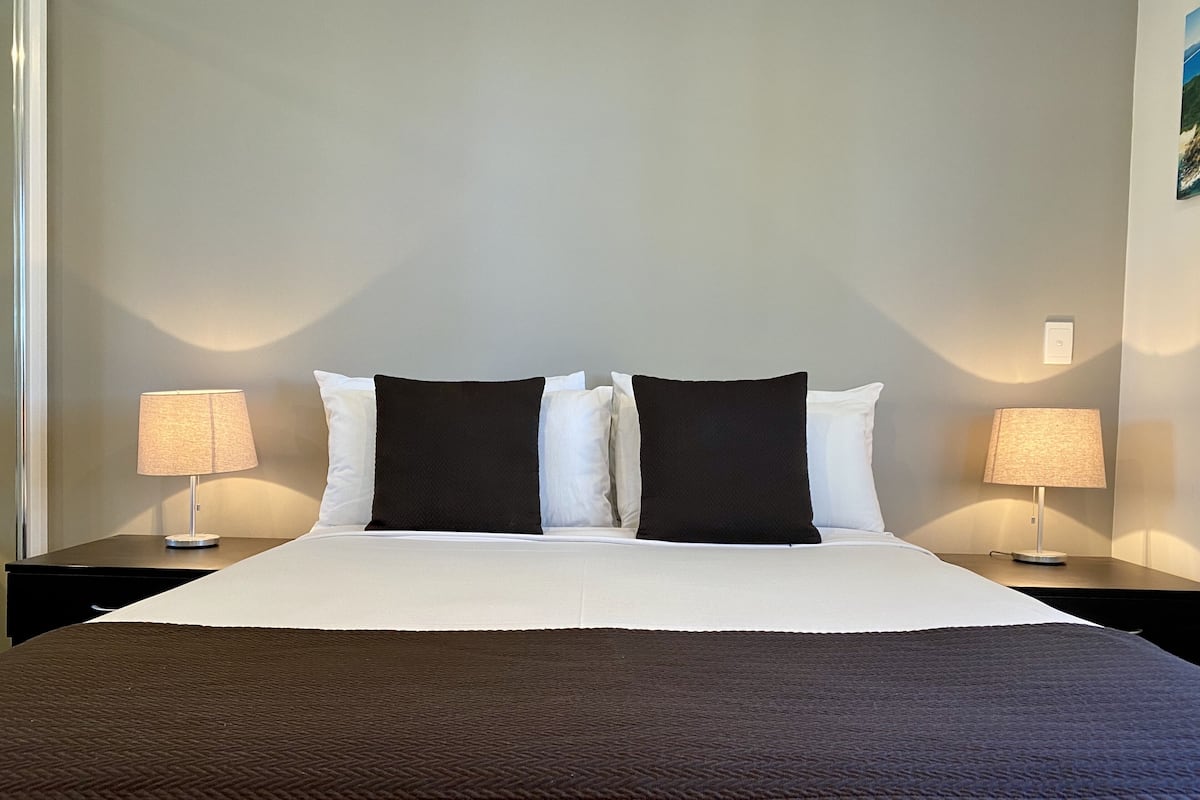 A neatly made bed with a white duvet and two black pillows, flanked by two bedside tables with lamps, against a plain wall.