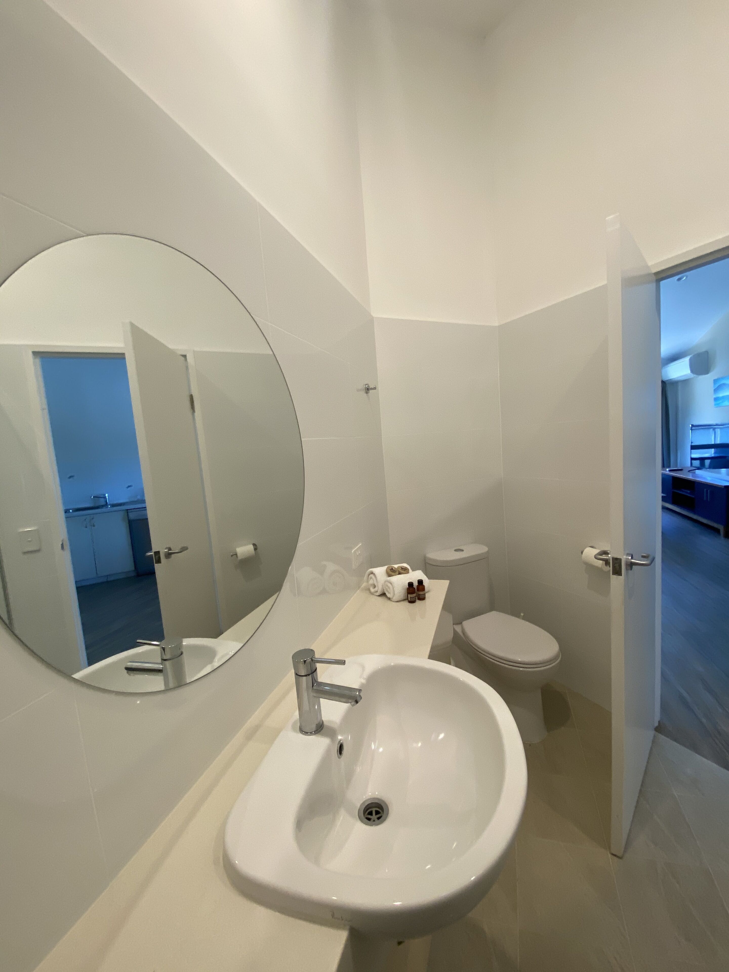 A modern bathroom with a large mirror, a sink, and a toilet. There is a towel rack with towels and a small bottle on the countertop.
