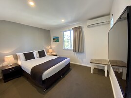Un dormitorio con cama, mesitas de noche, una ventana con cortinas, una pequeña mesa y un equipo de aire acondicionado.