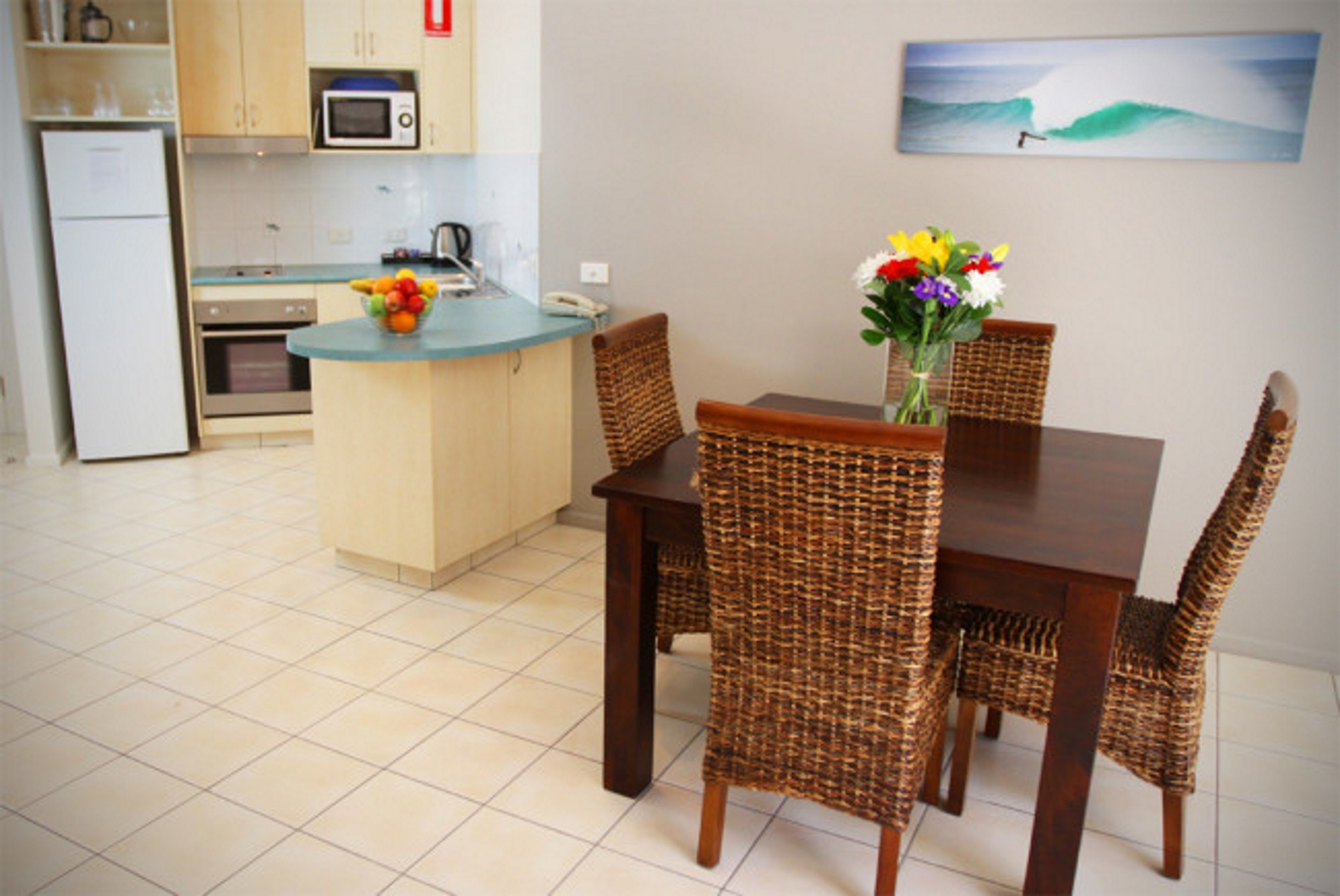 A kitchen with a dining area, featuring a small island with a fruit bowl, wicker chairs, and a vase with flowers.