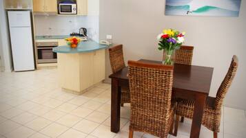 A kitchen with a dining area, featuring a small island with a fruit bowl, wicker chairs, and a vase with flowers.