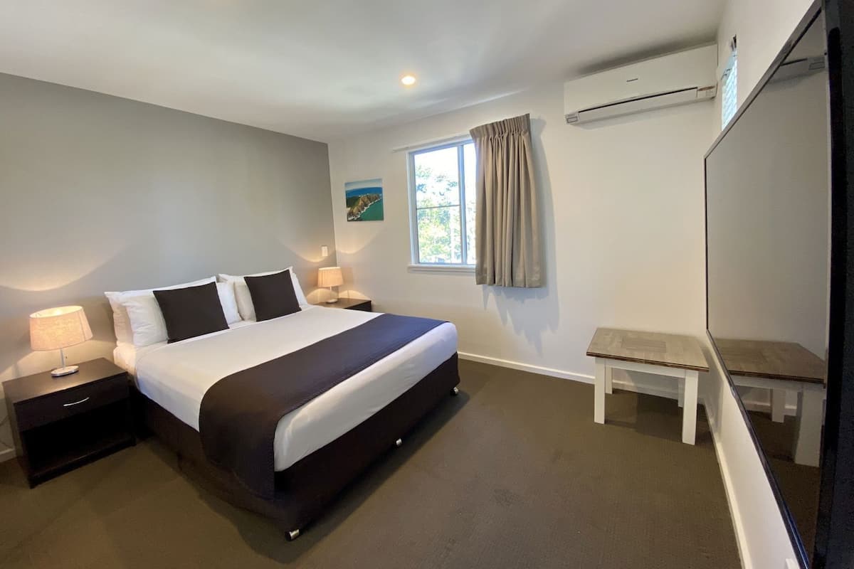 A bedroom with a bed, bedside tables, a window with curtains, a small table, and an air conditioning unit.