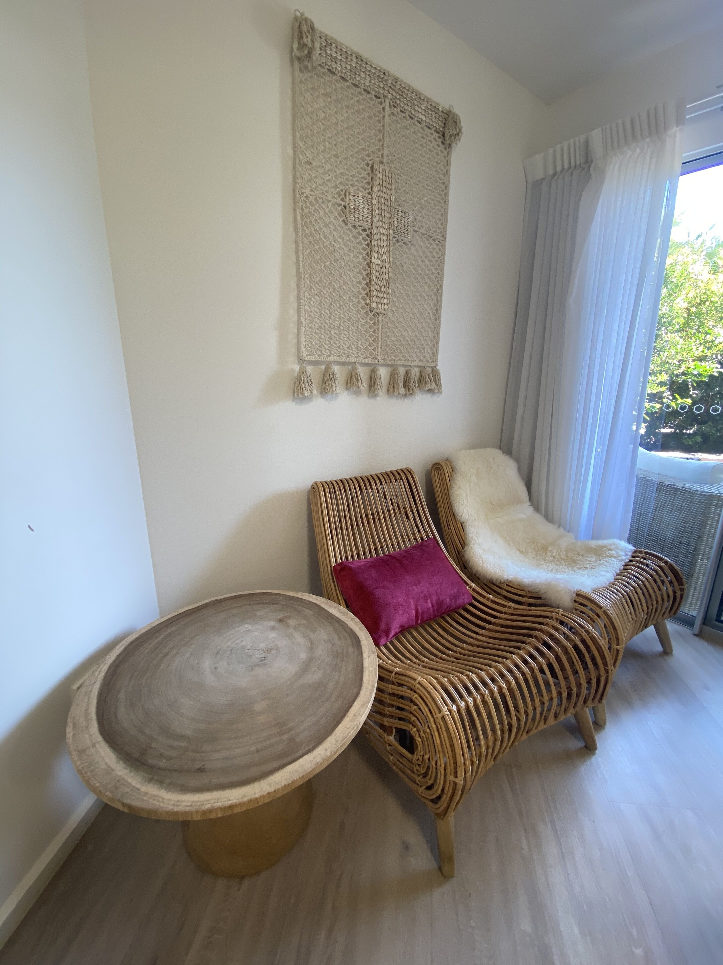 Un canapé en osier agrémenté d’un plaid blanc moelleux et d’un coussin violet, une décoration murale tissée et une table ronde en bois.