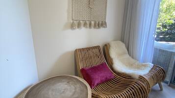 A wicker sofa with a fluffy white throw and a purple pillow, a woven wall hanging, and a round wooden table.