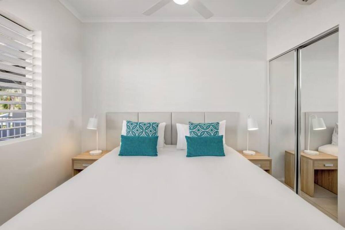 A bedroom with a bed, two bedside tables, a mirror, and a window with blinds.