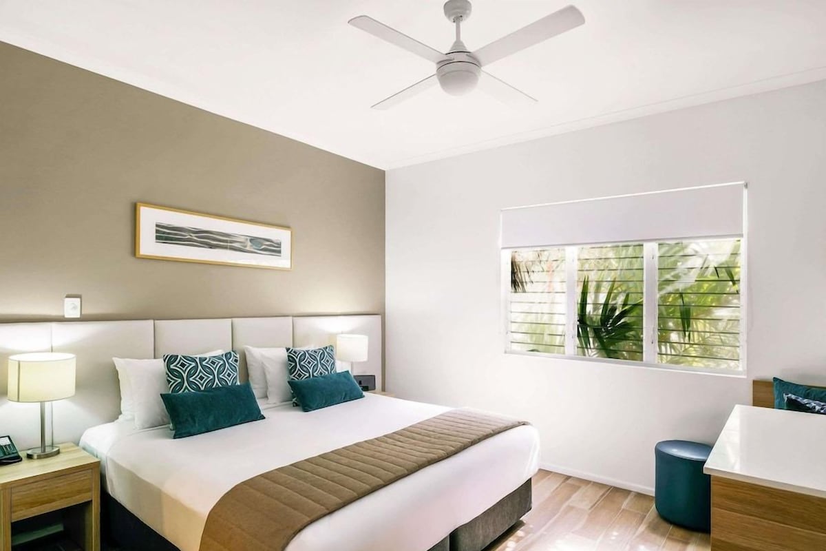 A modern hotel room with a large bed, a ceiling fan, a window with blinds, a bedside table with a lamp, and a small trash bin.