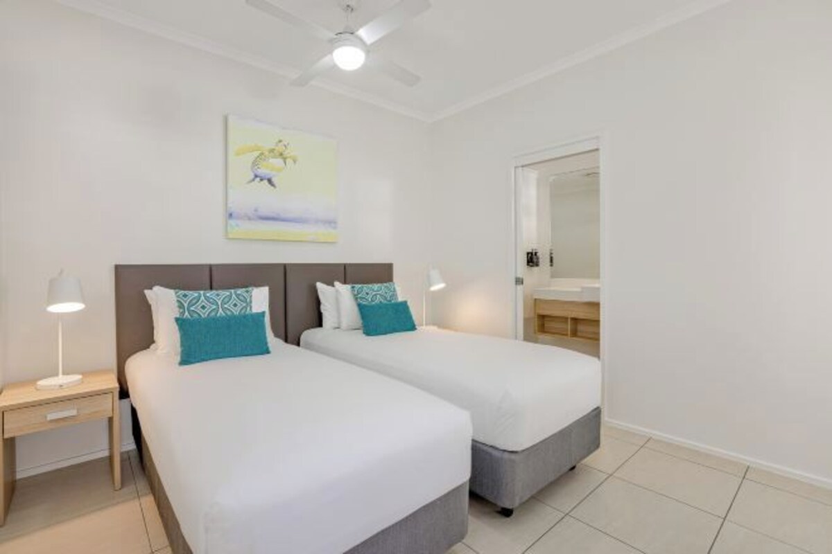 A hotel room with two beds, a ceiling fan, a painting, a nightstand, and a bathroom visible through an open door.