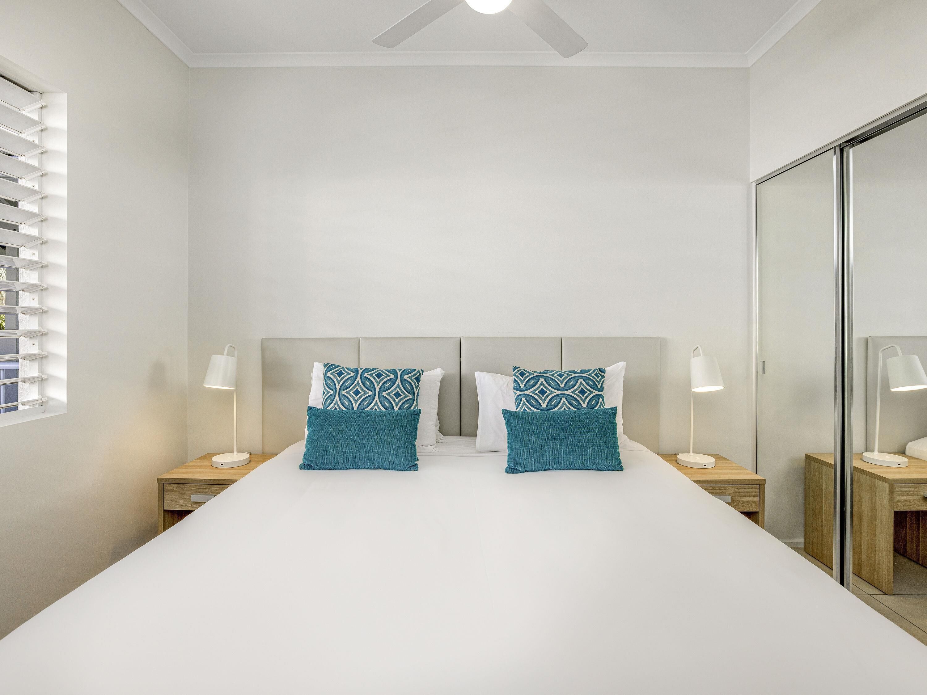 Une chambre avec un grand lit, deux tables de chevet avec des lampes, un ventilateur de plafond et un grand miroir.