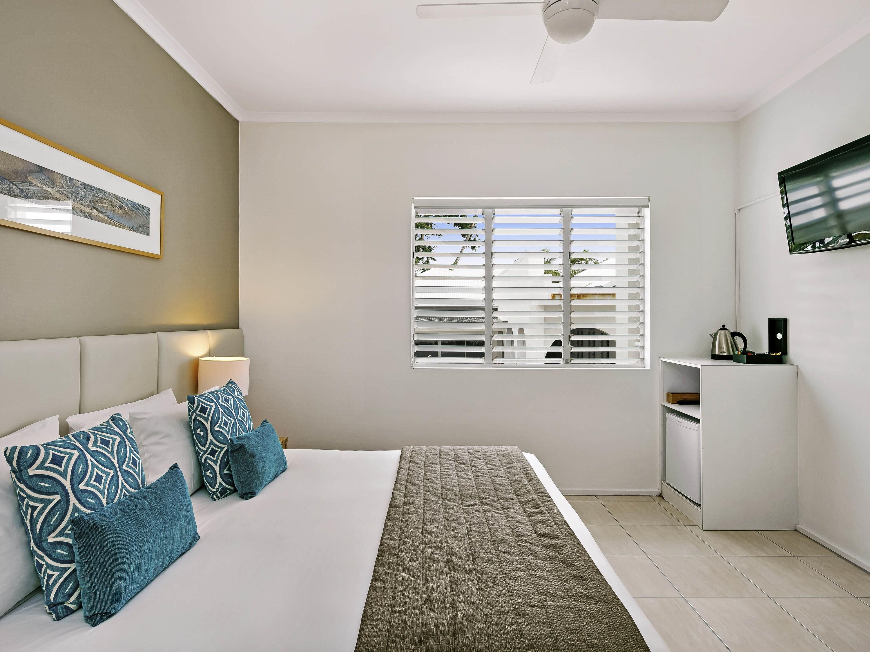 A modern hotel room with a bed, pillows, a bedside table, a lamp, a window with blinds, and a television mounted on the wall.