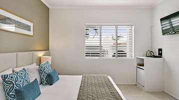 A modern hotel room with a bed, pillows, a bedside table, a lamp, a window with blinds, and a television mounted on the wall.