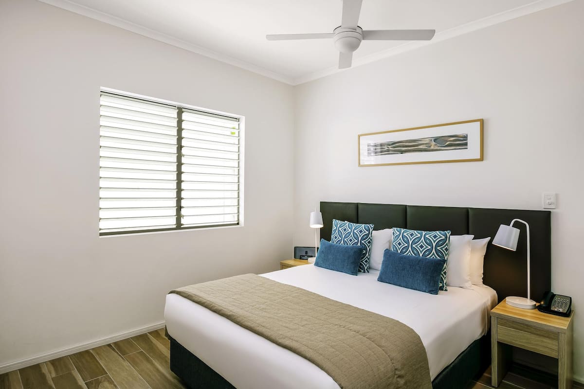 A bedroom with a bed, a ceiling fan, a window with blinds, a bedside table with a lamp, and a framed picture on the wall.