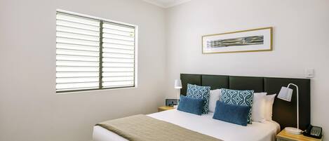 A bedroom with a bed, a ceiling fan, a window with blinds, a bedside table with a lamp, and a framed picture on the wall.