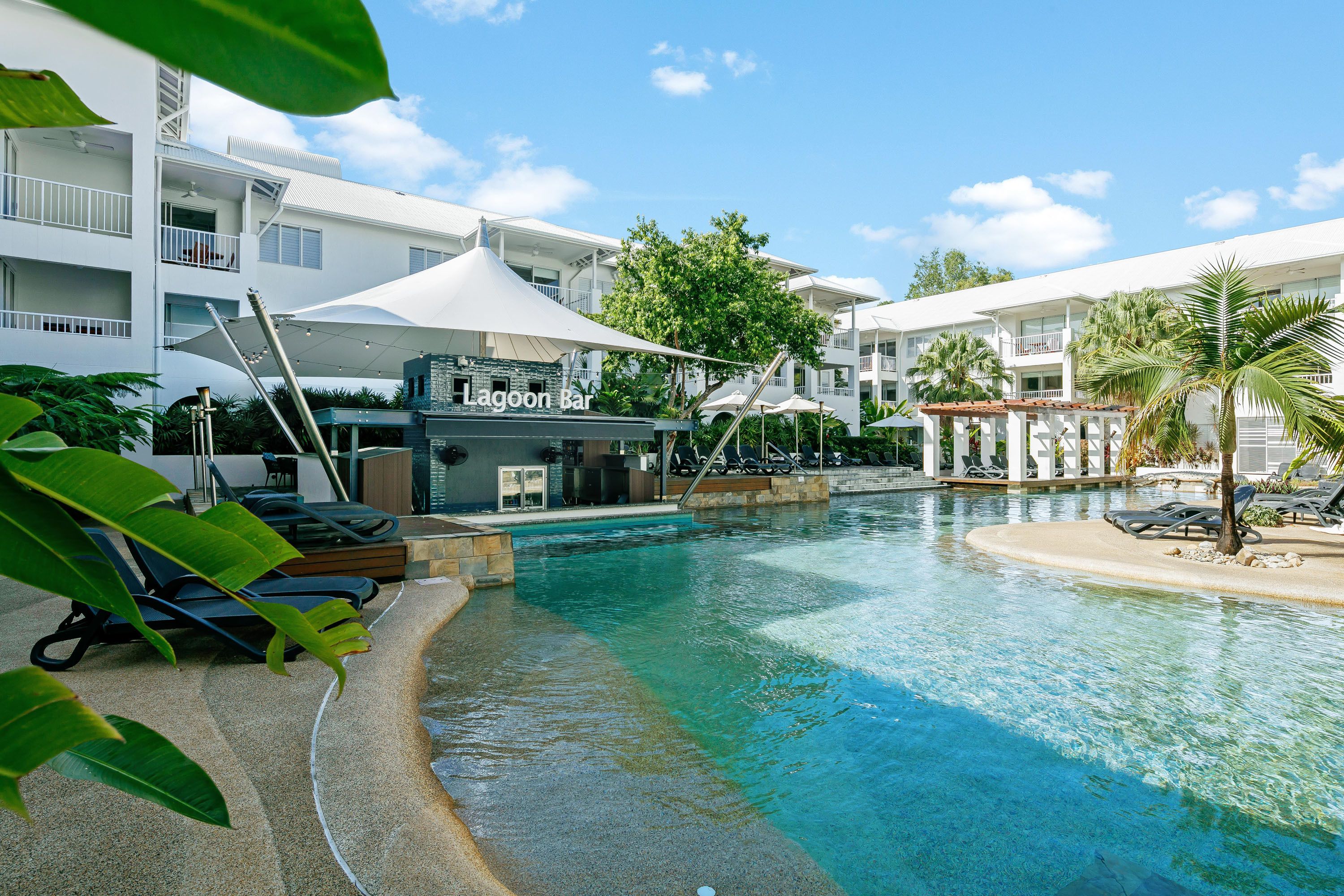 A swimming pool area with lounge chairs, a bar named 'Lagoon Bar', and a building with balconies.