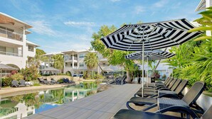 A poolside area with lounge chairs and umbrellas, a clear pool, and multi-story buildings in the background. - Mantra PortSea (Port Douglas)