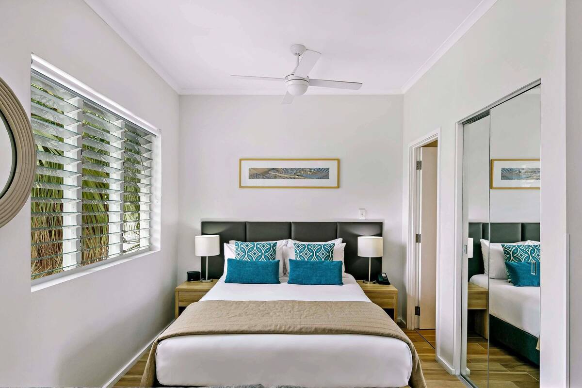 A hotel room with a bed, two bedside tables, a ceiling fan, a large window with blinds, and a mirror.