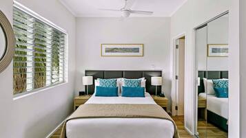 Une chambre d’hôtel comprenant un lit, deux tables de chevet, un ventilateur de plafond, une grande fenêtre avec des stores et un miroir.