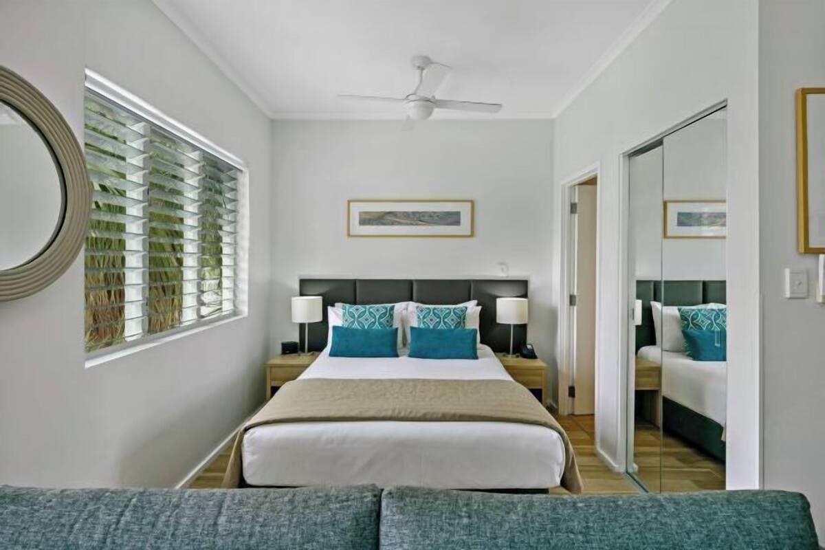 A hotel room with a bed, bedside tables, a ceiling fan, a window with blinds, and a framed picture on the wall.