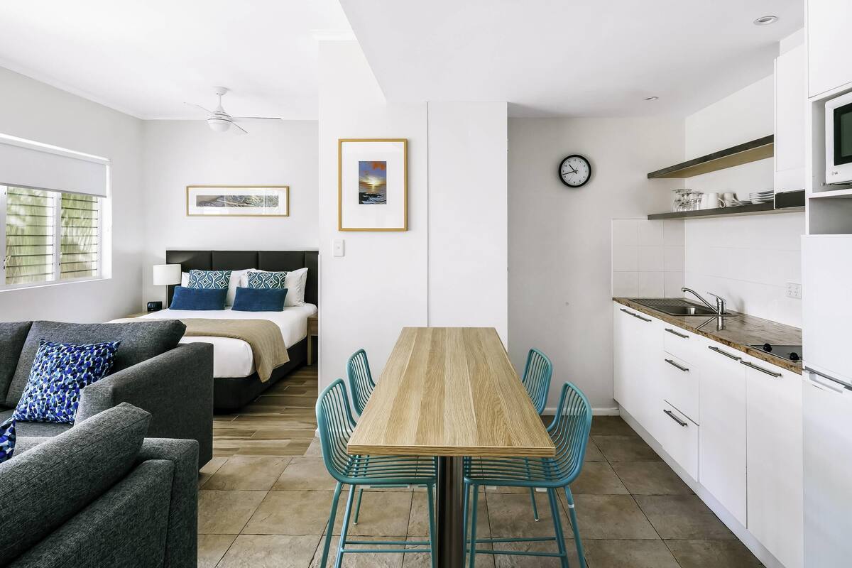 A modern, well-lit room with a bed, a sofa, a dining table with chairs, a kitchen area, and a clock on the wall.