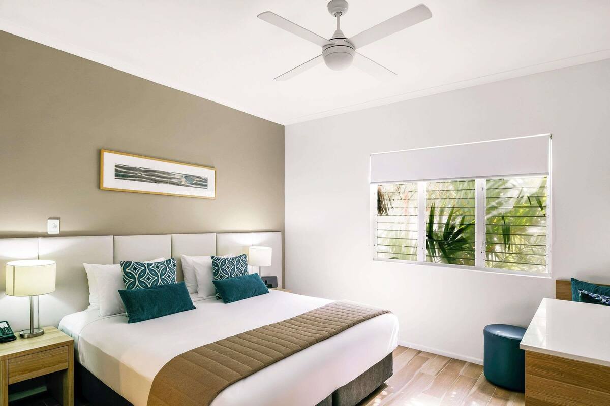 A hotel room with a bed, a ceiling fan, a window with blinds, a bedside table with a lamp, and a framed picture on the wall.