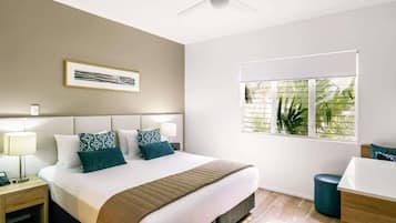 Una habitación de hotel con cama, ventilador de techo, ventana con persianas, mesita de noche con lámpara y un cuadro en la pared.