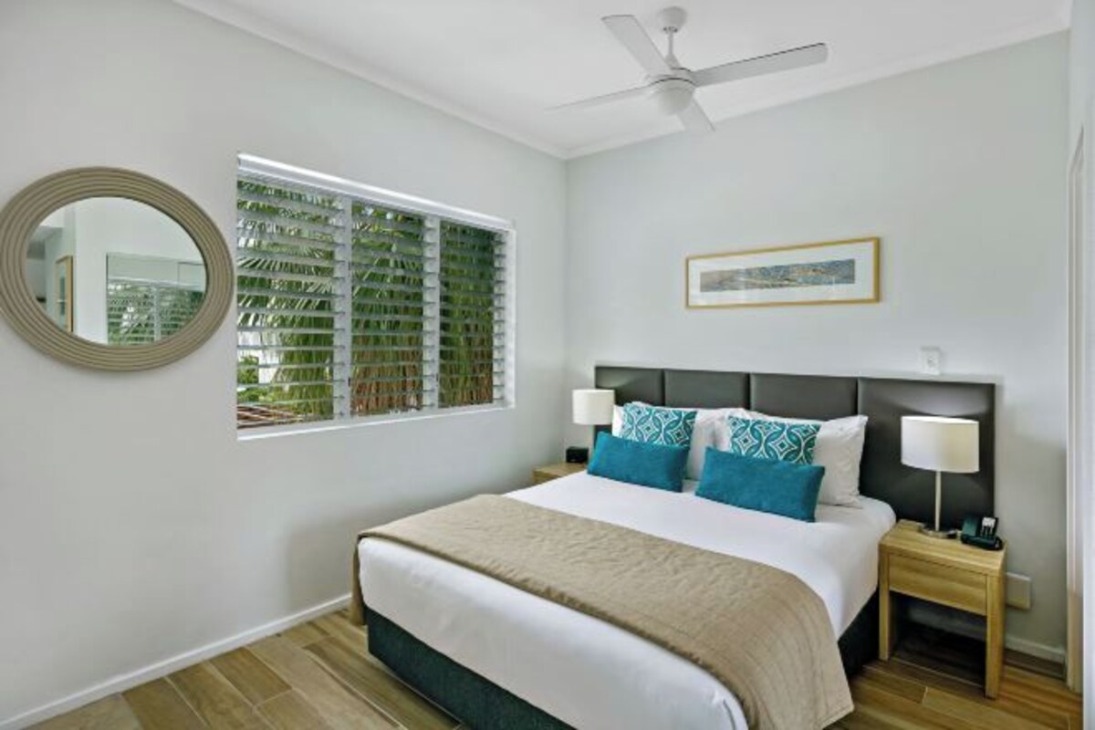 A bedroom with a bed, two bedside tables, a ceiling fan, a window with blinds, a framed picture on the wall, and a mirror.