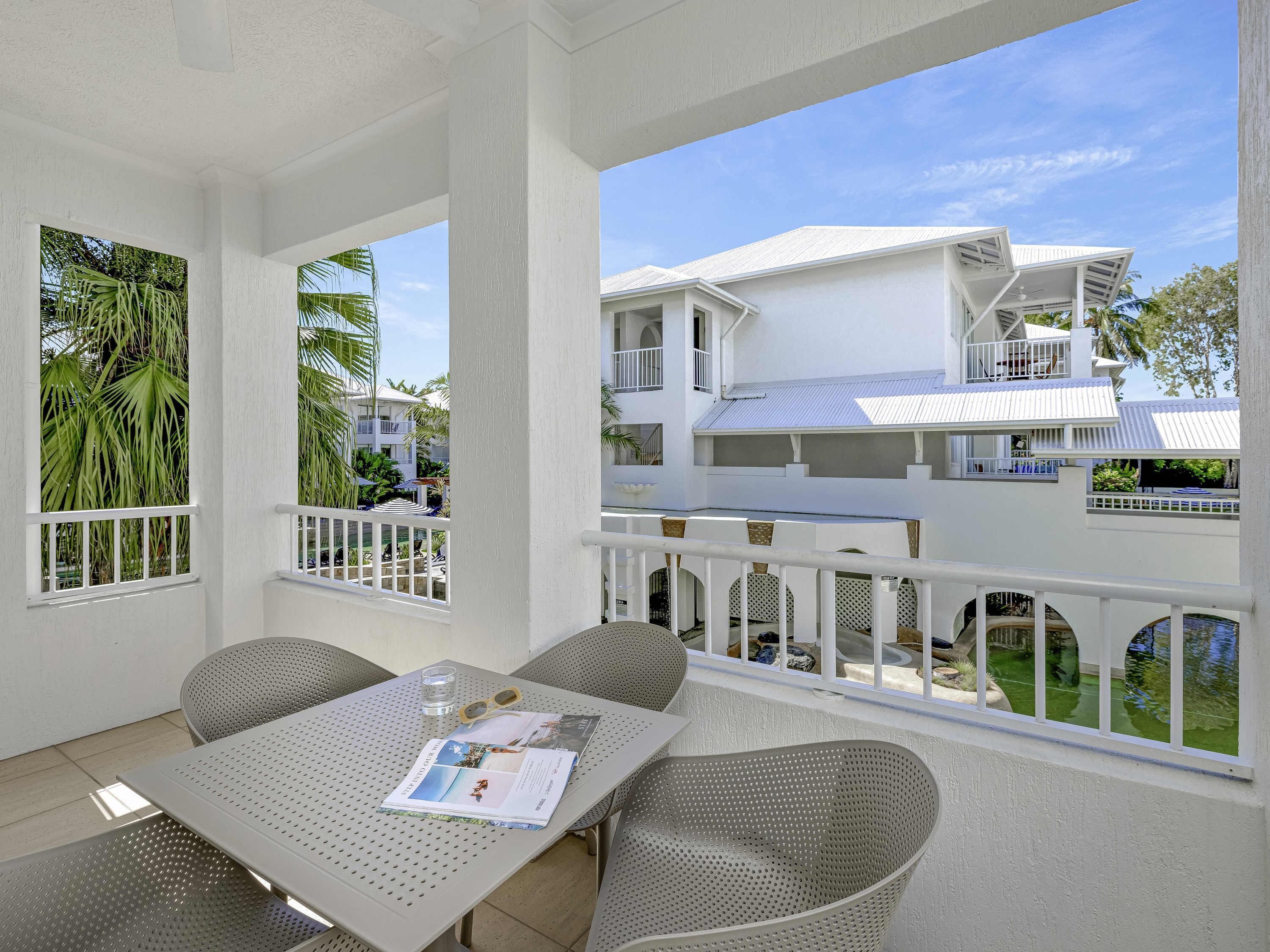 A covered outdoor seating area with a table, chairs, and a view of a white building with balconies.