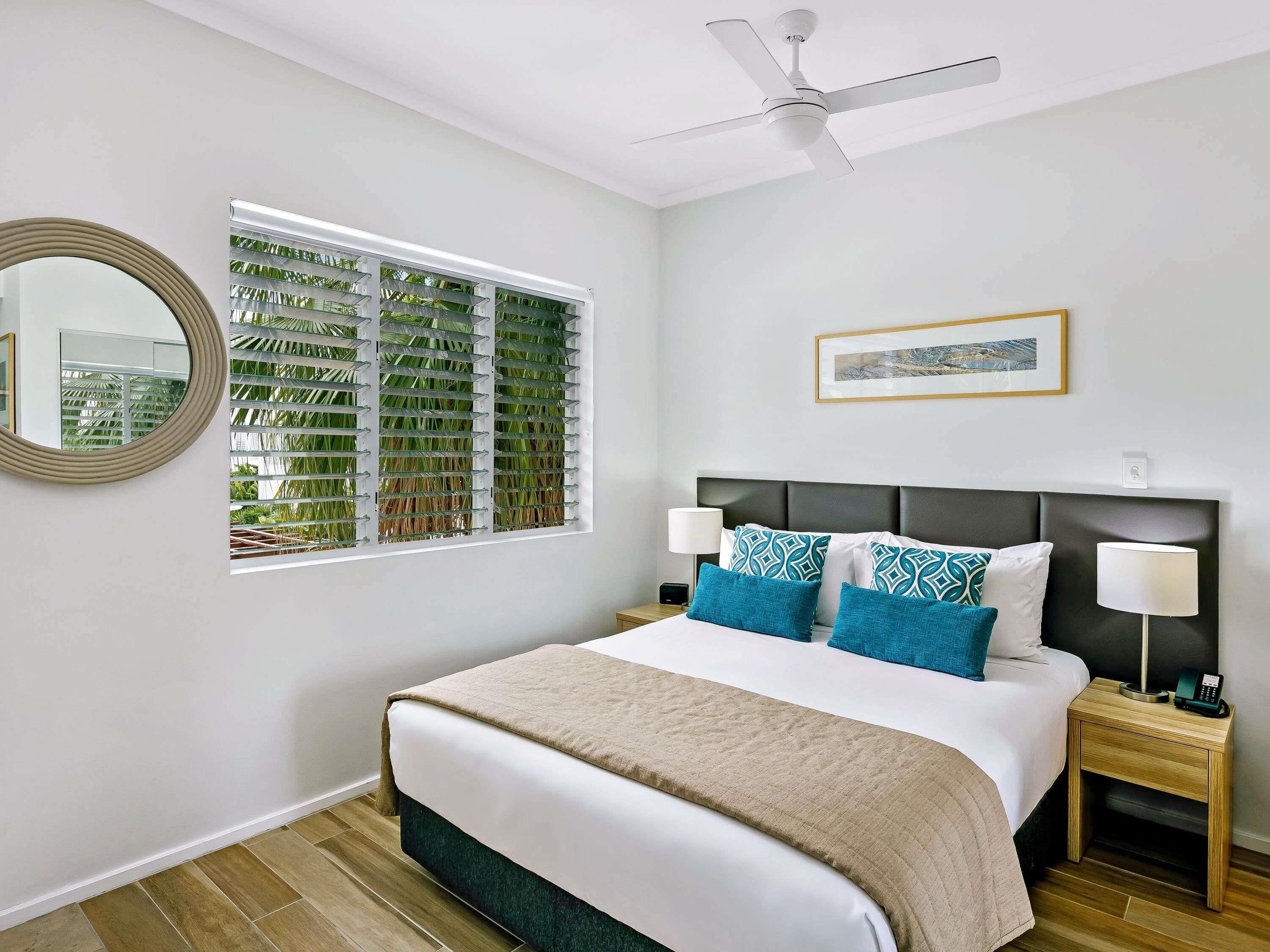 A bedroom with a bed, two bedside tables, a ceiling fan, a window with blinds, a framed picture, and a mirror.