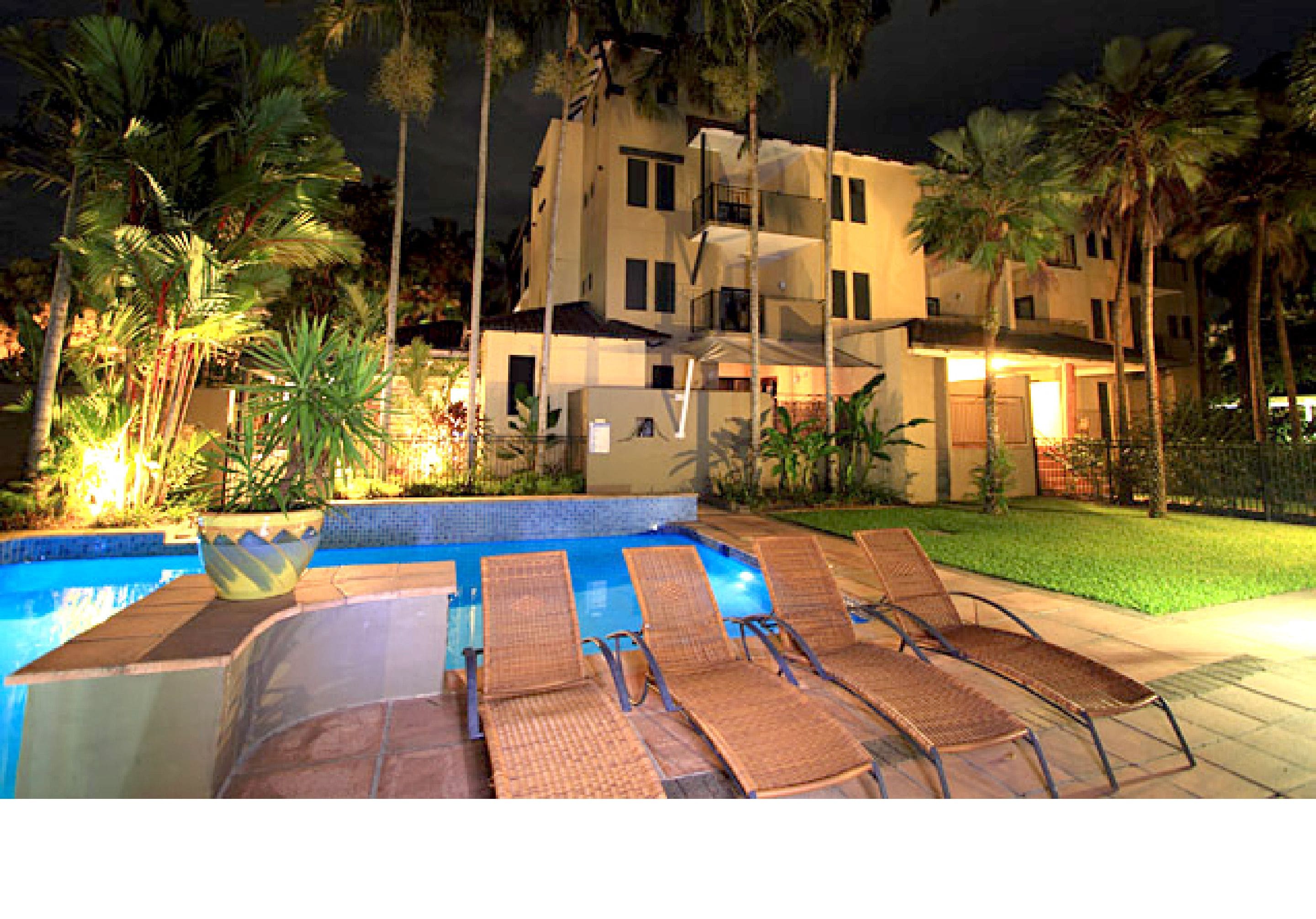 A swimming pool with lounge chairs, palm trees, and a multi-story building with balconies.