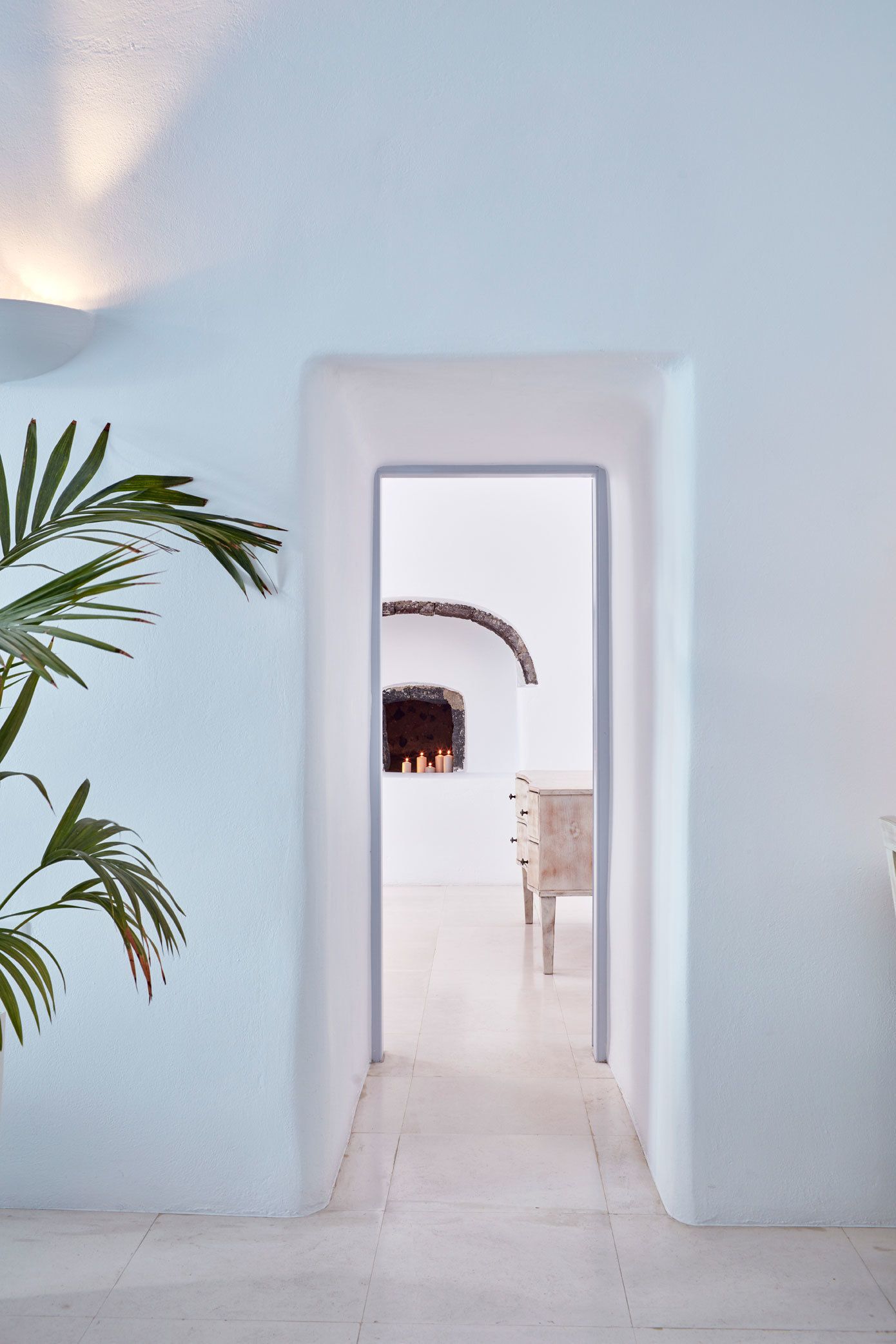 A hallway with a white wall, a plant on the left, and a doorway leading to a room with a fireplace.