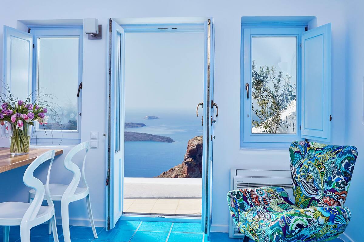 A room with a view of the sea, a white chair, a colorful chair, and a vase of flowers.