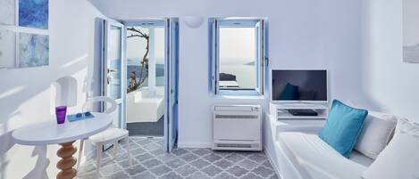 A modern, minimalist room with a white sofa, a small round table, and a TV. There are two open doors leading to a balcony with a view of the sea.