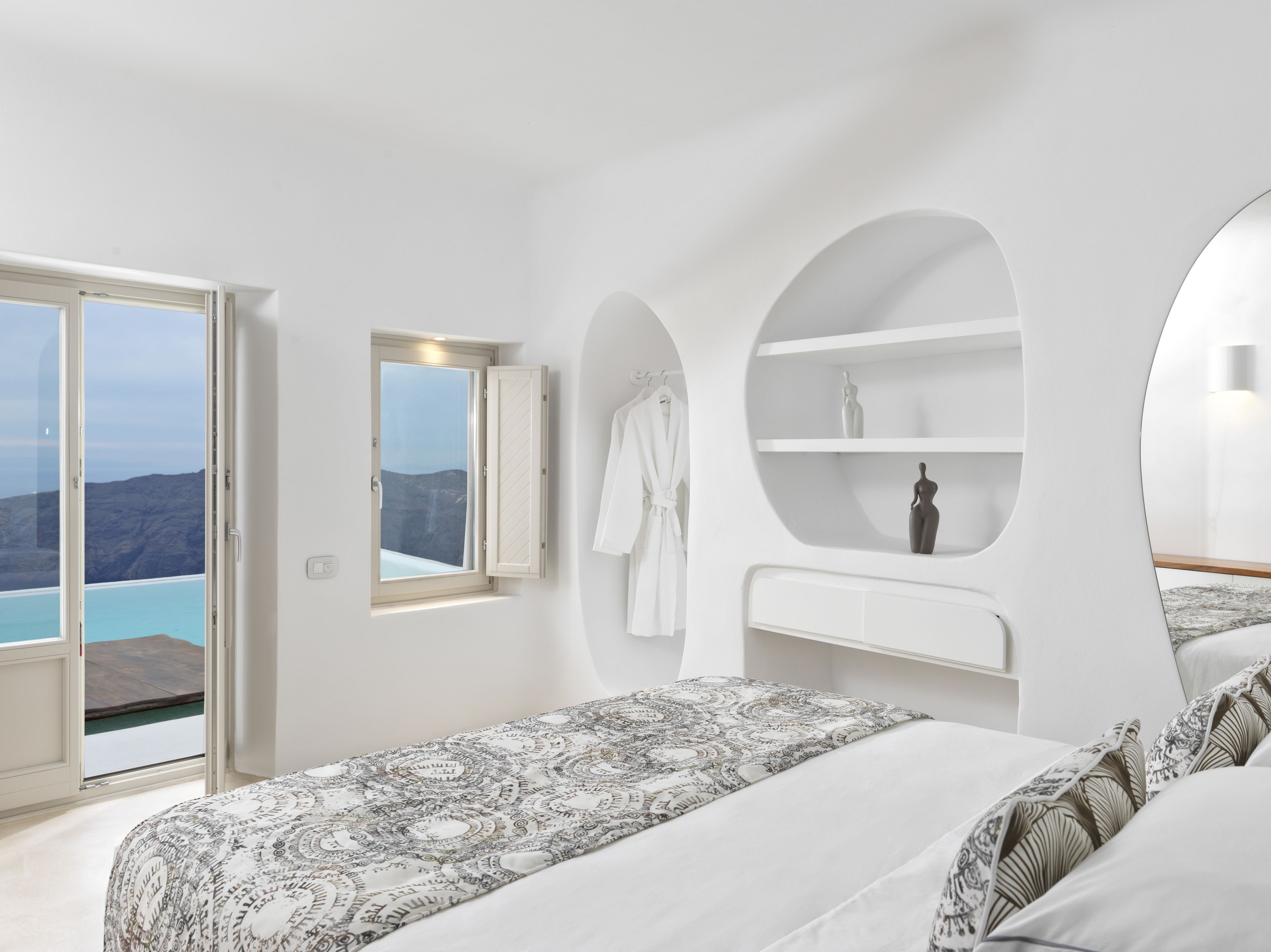 A modern bedroom with a large bed, a built-in shelf, a robe, and a view of the sea.