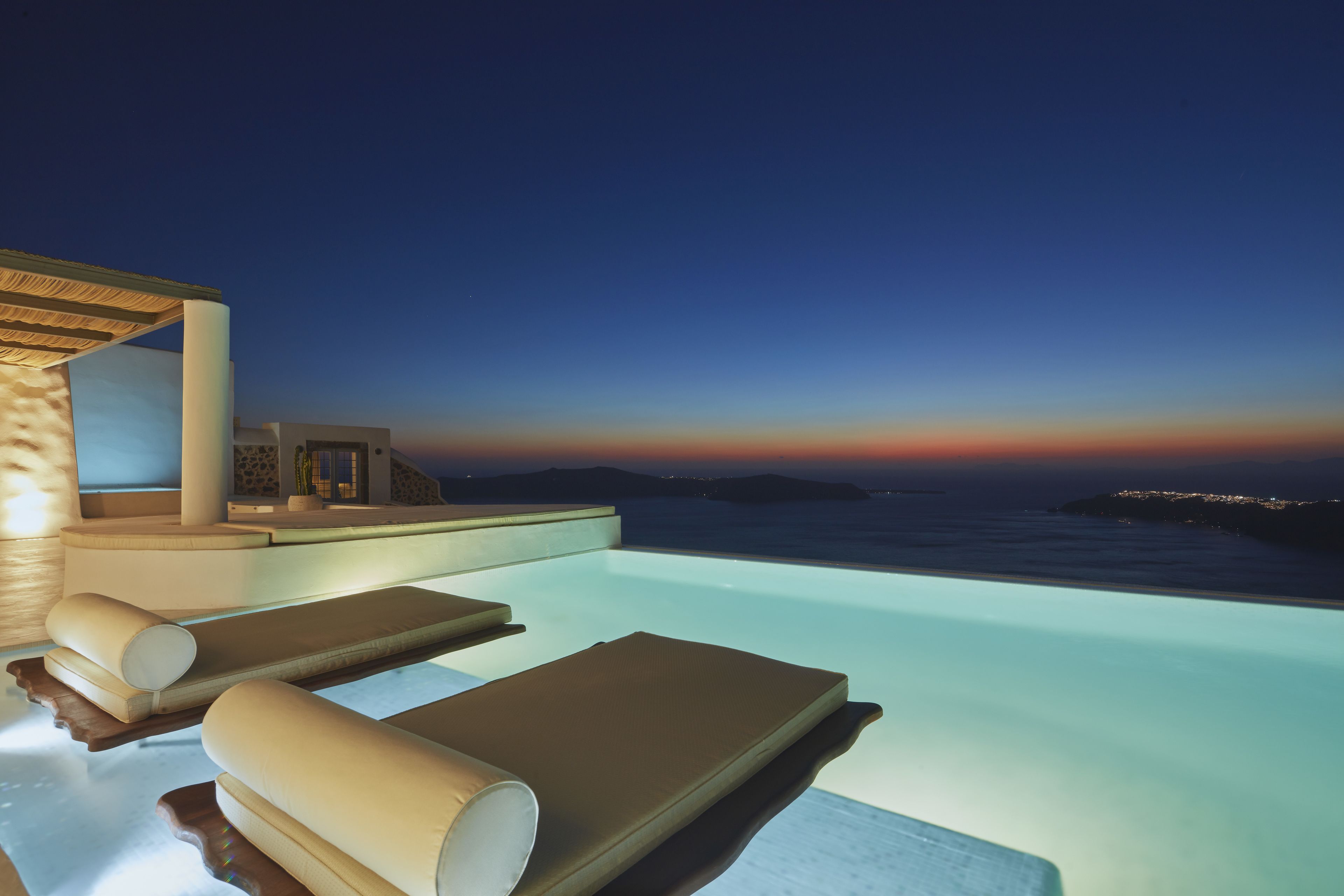 An infinity pool with lounge chairs, a pergola, and a view of the ocean at dusk.